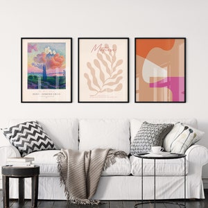 May include: Three framed art prints on a white wall above a white sofa. The left print is a landscape painting with a pink cloud and the text "Henri Edmond Cross". The middle print is a beige leaf with the text "Matisse". The right print is an abstract design with orange, pink, and beige shapes.