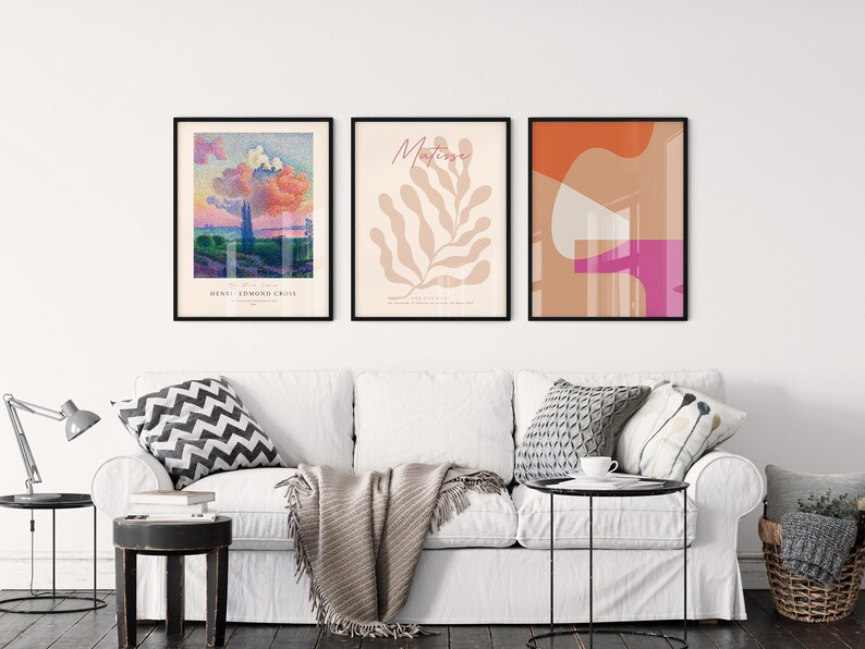 May include: Three framed art prints on a white wall above a white sofa. The left print is a landscape painting with a pink cloud and the text "Henri Edmond Cross". The middle print is a beige leaf with the text "Matisse". The right print is an abstract design with orange, pink, and beige shapes.
