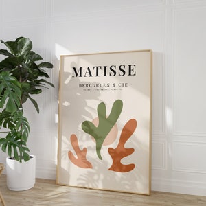 May include: A beige poster with a gold frame featuring a Matisse art print. The poster has a green, orange, and pink abstract design with the text "MATISSE" and "BERGGRUEN & CIE 70, RUE DE L'UNIVERSITE, PARIS-VII" in black.