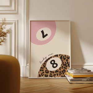 May include: A white framed print with a pink and beige background. The print features two 8-ball designs, one with the number 7 and the other with the number 8. The 8-ball with the number 8 is surrounded by a leopard print pattern. The text "lucky me" is written in cursive above the 8-ball.