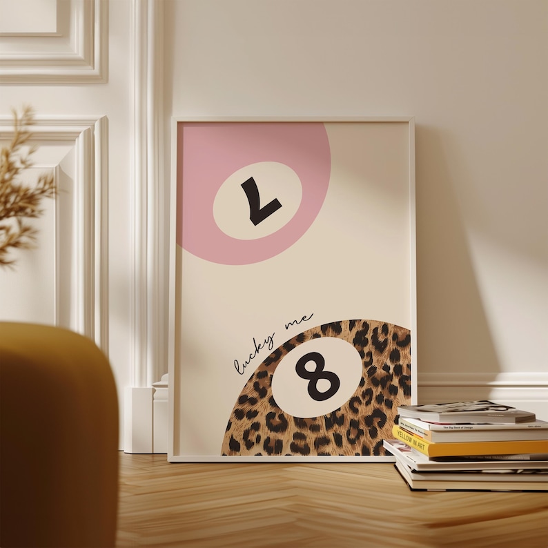 May include: A white framed print with a pink and beige background. The print features two 8-ball designs, one with the number 7 and the other with the number 8. The 8-ball with the number 8 is surrounded by a leopard print pattern. The text "lucky me" is written in cursive above the 8-ball.
