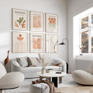 May include: A living room interior with a white couch, two armchairs, a coffee table, and a floor lamp. The wall is decorated with six framed prints featuring abstract art in shades of brown, orange, and green. The prints have text that reads "Matisse" and "Flower Market".