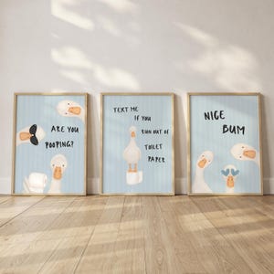 May include: Three framed wall art prints featuring cartoon geese on a light blue striped background. The first print says ARE YOU POOPING?, the second says TEXT ME IF YOU RUN OUT OF TOILET PAPER, and the third says NICE BUM with goose illustrations.