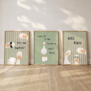 May include: Three framed wall art prints featuring white geese on sage green backgrounds. The first print has pink stripes and text reading ARE YOU POOPING?. The second print shows a goose on toilet paper with text reading TEXT ME IF YOU RUN OUT OF TOILET PAPER. The third print has pink stripes and text reading NICE BUM.