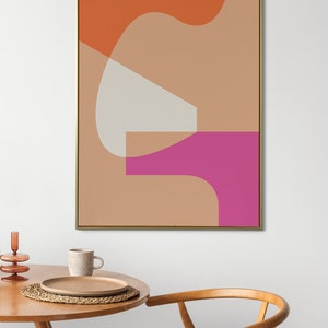 May include: Abstract art print featuring geometric shapes in orange, pink, and beige colours. The print is framed in gold and hangs on a white wall above a wooden table and chair.