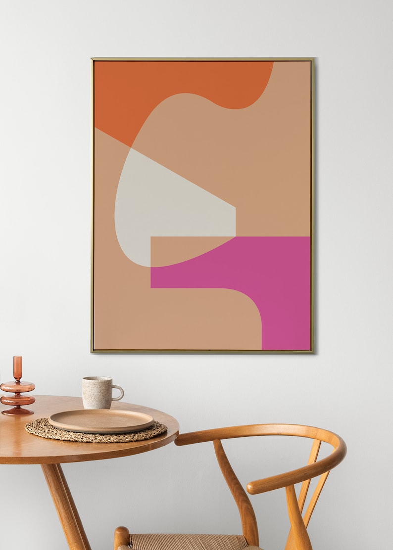 May include: Abstract art print featuring geometric shapes in orange, pink, and beige colours. The print is framed in gold and hangs on a white wall above a wooden table and chair.