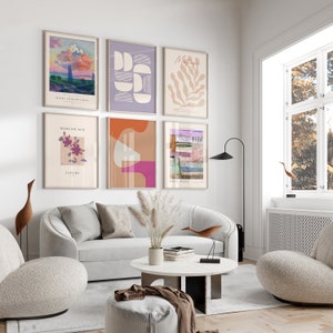 May include: A living room interior featuring a white sofa, two armchairs, a coffee table, and a floor lamp. The wall is adorned with six framed art prints, showcasing abstract geometric shapes, a floral design, and a landscape scene. The prints are in a variety of colours, including pink, orange, purple, and green.