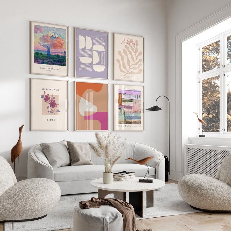 May include: A living room interior featuring a white sofa, two armchairs, a coffee table, and a floor lamp. The wall is adorned with six framed art prints, showcasing abstract geometric shapes, a floral design, and a landscape scene. The prints are in a variety of colours, including pink, orange, purple, and green.