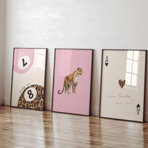 May include: Three framed prints with a pink, cream, and white background. The left print features a pink circle with the letter "L" and a leopard print 8 ball with the text "lucky me". The middle print features a pink background with a leopard print animal. The right print features a cream background with a black ace of spades with a red heart and the text "how lucky are we?"