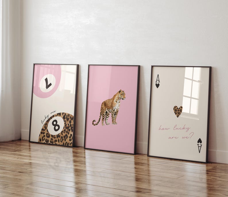 May include: Three framed prints with a pink, cream, and white background. The left print features a pink circle with the letter "L" and a leopard print 8 ball with the text "lucky me". The middle print features a pink background with a leopard print animal. The right print features a cream background with a black ace of spades with a red heart and the text "how lucky are we?"