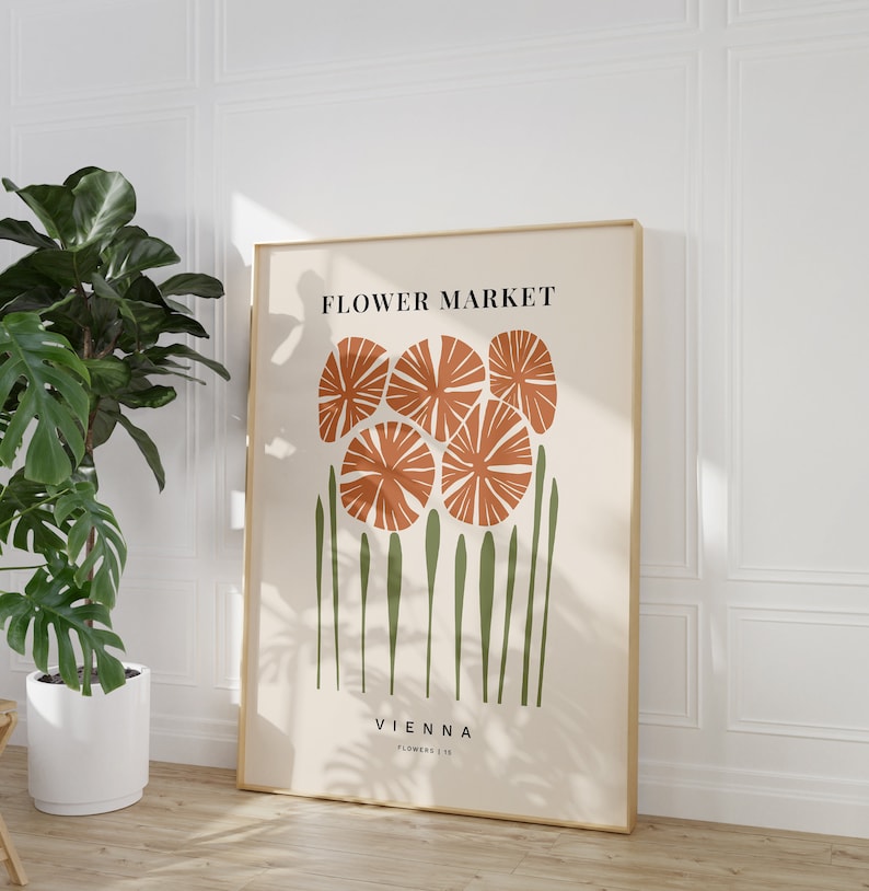 May include: A beige poster with a gold frame features a stylized illustration of orange flowers with green stems. The text "FLOWER MARKET" and "VIENNA" is printed in black.