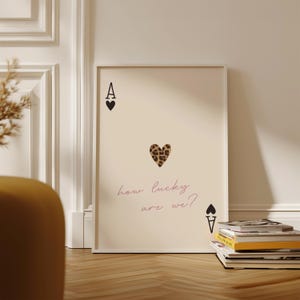 May include: A beige poster featuring a black ace of spades playing card with a leopard print heart in the centre. The text "how lucky are we?" is written in pink below the heart.