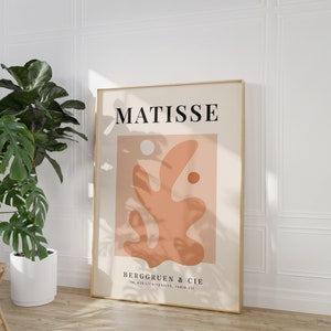 May include: A minimalist art print featuring a stylized abstract design in shades of peach and beige. The print is framed in gold and includes the text "MATISSE" and "BERGGRUEN & CIE 70, RUE DE L'UNIVERSITE, PARIS-VII".