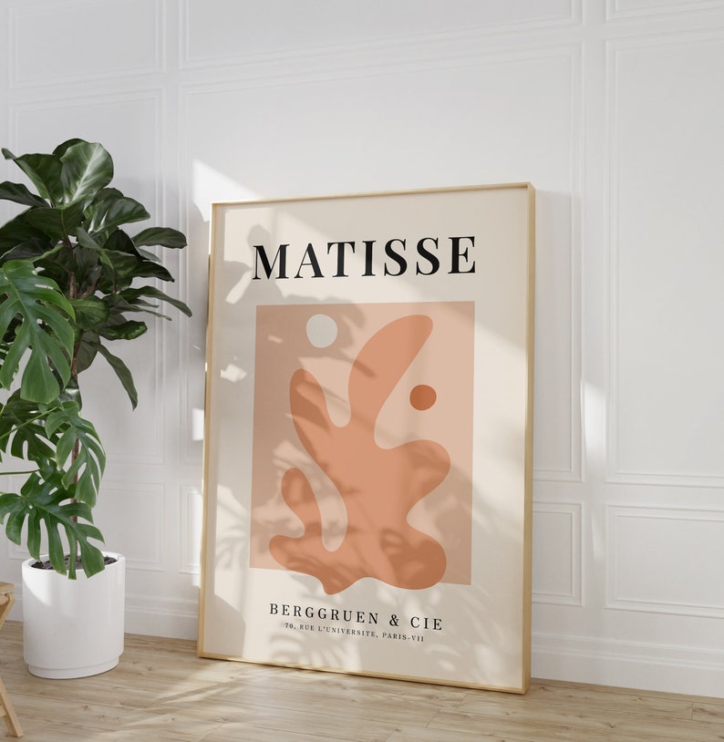 May include: A minimalist art print featuring a stylized abstract design in shades of peach and beige. The print is framed in gold and includes the text "MATISSE" and "BERGGRUEN & CIE 70, RUE DE L'UNIVERSITE, PARIS-VII".