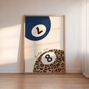 Op de afbeelding: Een ingelijste print met een witte achtergrond en twee biljartballen. De bovenste bal is blauw met het nummer 1 in wit. De onderste bal is bruin met een luipaardprint en het nummer 8 in zwart. De tekst "lucky me" is geschreven in zwarte cursieve letters.
