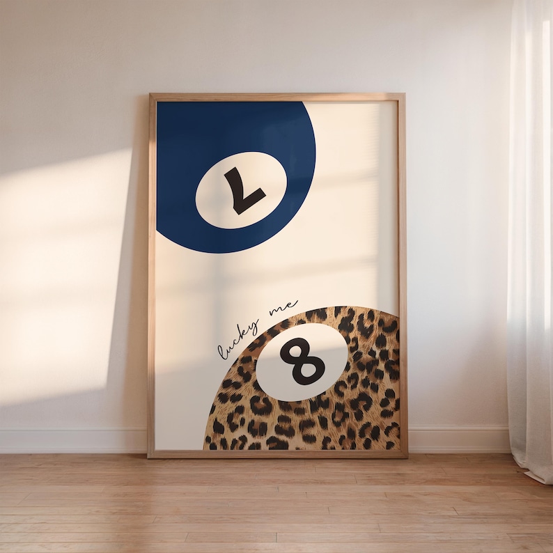 Op de afbeelding: Een ingelijste print met een witte achtergrond en twee biljartballen. De bovenste bal is blauw met het nummer 1 in wit. De onderste bal is bruin met een luipaardprint en het nummer 8 in zwart. De tekst "lucky me" is geschreven in zwarte cursieve letters.
