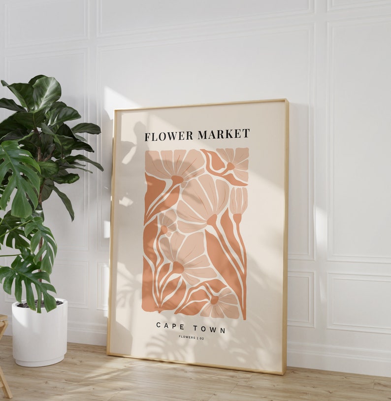 May include: A beige poster with a gold frame featuring a stylized illustration of pink and orange flowers. The text "FLOWER MARKET" and "CAPE TOWN" is printed in black on the poster.
