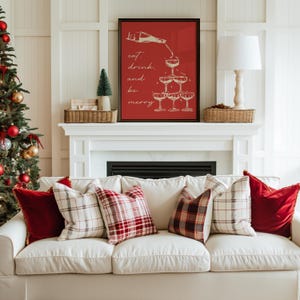K&ouml;nnte beinhalten: Ein wei&szlig;es Sofa mit roten und schwarzen karierten Kissen in einem Wohnzimmer mit einem Kamin und einem Weihnachtsbaum. Ein gerahmtes Bild &uuml;ber dem Kamin zeigt den Text "eat, drink, and be merry" mit einer Zeichnung einer Champagnerflasche, die in eine Pyramide von Champagnergl&auml;sern gegossen wird.
