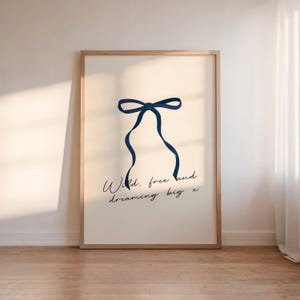 Op de afbeelding: Een beige poster met een blauw lint dat in een strik is gebonden. De tekst "Wild. free and dreaming big x" is geschreven in zwarte cursieve letters.