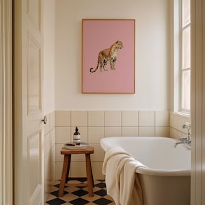 May include: A white bathtub with a white towel on the side, a wooden stool with a bottle of soap and a tray on top, and a framed print of a leopard on a pink background hanging on the wall above the bathtub.