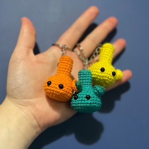May include: Three crocheted keychains in the shape of a small, round, bulbous object with two black eyes. The keychains are orange, yellow, and teal.