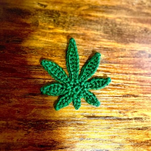 May include: A green crocheted marijuana leaf on a wooden surface.