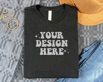 May include: A black t-shirt with the text "YOUR DESIGN HERE" in a retro font. The shirt is folded and laid on a gray and white patterned fabric.