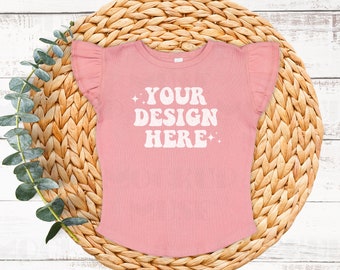 May include: A pink ruffle-sleeved t-shirt with the text "YOUR DESIGN HERE" in white. The shirt is on a woven straw placemat.