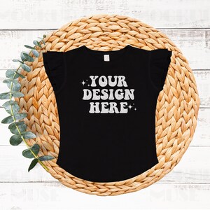 Puede incluir: Camiseta negra con ribete de volantes en las mangas. La camiseta tiene el texto "YOUR DESIGN HERE" impreso en la parte delantera en una fuente retro blanca.