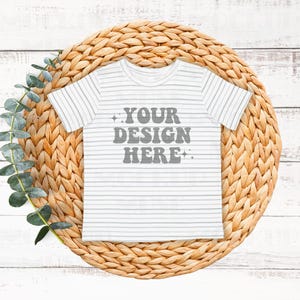 May include: White and gray striped baby t-shirt with the text "YOUR DESIGN HERE" in gray on a woven straw mat.
