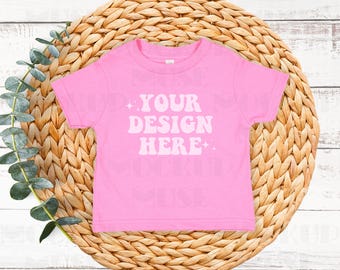 May include: Pink baby t-shirt with the text "YOUR DESIGN HERE" in white retro font on a woven straw background.