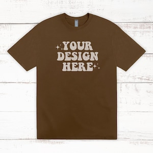 May include: A brown t-shirt with the text "YOUR DESIGN HERE" in a retro font. The short-sleeved shirt is a solid color and is displayed on a white wooden surface.