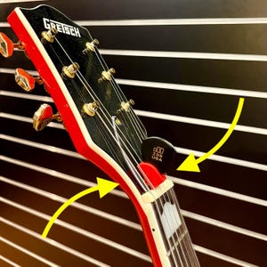 May include: Close-up of the headstock of a red and black electric guitar with gold tuning pegs. The guitar has a white binding around the edge of the headstock. A black guitar pick is attached to the headstock with the text "000 208 USA".