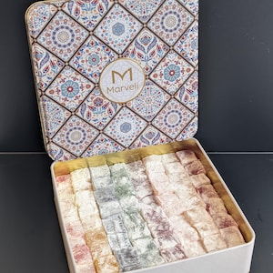 May include: A square metal tin filled with assorted colorful Turkish delight cubes. The lid features a geometric pattern in blue, red, and white with a central circular logo reading M Marveli. The base of the tin is white with a subtle diamond texture.
