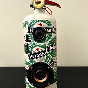 May include: A white fire extinguisher repurposed as a speaker, featuring multiple green and white Heineken brand logo stickers. The device includes two black circular speakers mounted on the front and a red handle with a pressure gauge on top.