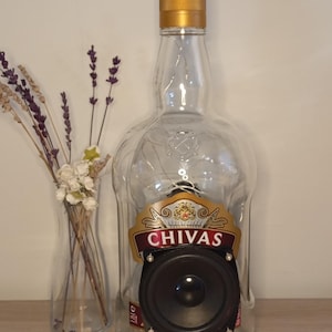 Può includere: Una bottiglia di whisky Chivas riutilizzata e trasformata in un altoparlante. La bottiglia di vetro trasparente presenta un tappo dorato e un altoparlante incorporato nella parte anteriore. L'etichetta Chivas è visibile. Una piccola candela e un vaso di fiori secchi sono vicini.