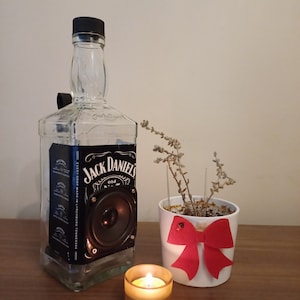 Può includere: Una bottiglia di vetro trasparente con un'etichetta nera che recita "Jack Daniel's Old No. 7 Tennessee Whiskey" è stata riadattata come altoparlante. La bottiglia è appoggiata su un tavolo di legno accanto a un vaso di ceramica bianca con un fiocco rosso e una piccola candela accesa.