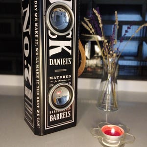 May include: A black and white Jack Daniel's whiskey barrel-shaped display with two circular mirrors. The text "Jack Daniel's" and "Handcrafted Barrels" are visible. A small glass vase with purple flowers and a flower-shaped candle holder with a lit candle are also present.