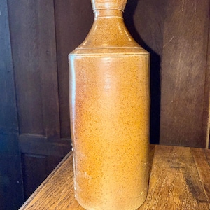 May include: A tall, cylindrical stoneware bottle with a speckled tan and brown finish. The bottle features a narrow neck with a flared rim and a smooth, rounded body. The surface has a textured, earthy appearance with subtle variations in color tone.