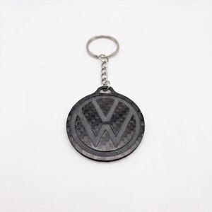 以下が含まれることがあります： フォルクスワーゲンのロゴが入った黒とグレーのキーホルダー。丸いペンダントはカーボンファイバーの質感で、シルバーのチェーンとキーリングに付いています。VWエンブレムが中央に大きく表示されています。
