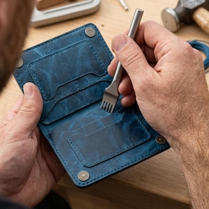 May include: A blue leather wallet with a distressed finish featuring multiple card slots and silver-toned metal snaps. A metal stitching chisel is being used to punch holes into the leather surface of the wallet during the assembly process.