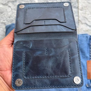 May include: A dark blue bi-fold wallet with a distressed texture, featuring multiple card slots and two silver-toned magnetic snap closures at the top and bottom corners. The wallet has visible stitching along the edges and interior pockets.