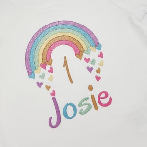 Puede incluir: Una camiseta blanca de manga corta con un dise&ntilde;o bordado de un arco&iacute;ris en tonos pastel con corazones peque&ntilde;os, el n&uacute;mero 1 y el nombre Josie en letras de colores. El cuello incluye dos botones a presi&oacute;n y una etiqueta de talla 12-18m.