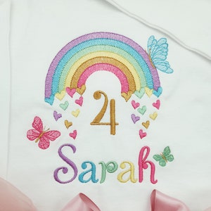 Könnte beinhalten: Ein weißes Kleidungsstück mit einer Stickerei aus einem bunten Regenbogen, kleinen Herzen und Schmetterlingen. Die Zahl 4 ist mittig in Goldfaden gestickt, darunter steht der Name Sarah in bunten Buchstaben. Eine rosa Tüllschleife ist angebracht.