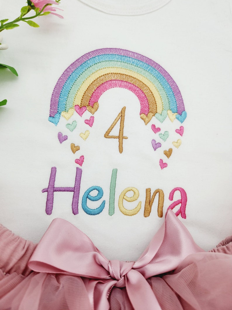 Puede incluir: Una camiseta blanca con un arco&iacute;ris bordado, corazones peque&ntilde;os, el n&uacute;mero 4 y el nombre Helena en hilo multicolor. Debajo, una falda de tul rosa empolvado con un lazo de cinta de sat&eacute;n a juego en el centro de la cintura.