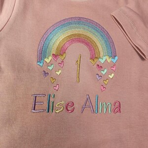 Puede incluir: Una camiseta de manga corta color rosa empolvado con un arco&iacute;ris bordado, corazones multicolores, el n&uacute;mero 1 y el nombre Elise Alma en hilo de colores. Una etiqueta blanca en el cuello muestra la talla 12-18m en texto negro.