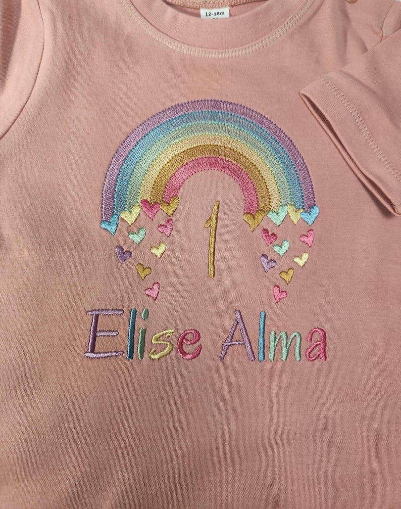 Puede incluir: Una camiseta de manga corta color rosa empolvado con un arco&iacute;ris bordado, corazones multicolores, el n&uacute;mero 1 y el nombre Elise Alma en hilo de colores. Una etiqueta blanca en el cuello muestra la talla 12-18m en texto negro.