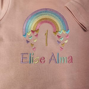 Puede incluir: Una camiseta de manga corta color rosa empolvado con un arco&iacute;ris bordado y peque&ntilde;os corazones cayendo. El n&uacute;mero 1 est&aacute; cosido en el centro y el nombre Elise Alma est&aacute; bordado debajo con hilo multicolor. Se ve una etiqueta de talla 12-18m.
