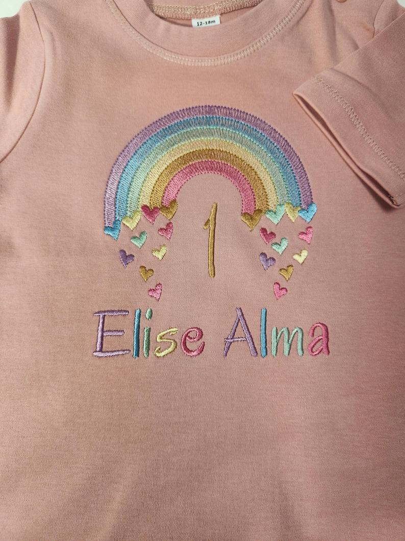 Puede incluir: Una camiseta de manga corta color rosa empolvado con un arco&iacute;ris bordado y peque&ntilde;os corazones cayendo. El n&uacute;mero 1 est&aacute; cosido en el centro y el nombre Elise Alma est&aacute; bordado debajo con hilo multicolor. Se ve una etiqueta de talla 12-18m.