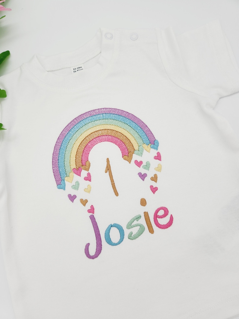Puede incluir: Una camiseta blanca de manga corta con un dise&ntilde;o bordado de un arco&iacute;ris en tonos pastel con corazones peque&ntilde;os, el n&uacute;mero 1 y el nombre Josie en letras de colores. El cuello incluye dos botones a presi&oacute;n y una etiqueta de talla 12-18m.